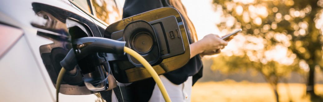 Eine Frau steht neben einem Elektroauto, das gerade aufgeladen wird und schaut auf ihr Smartphone.