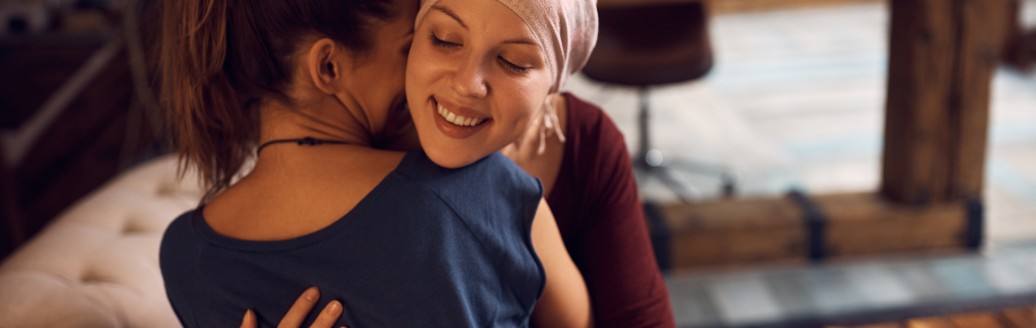 Zwei junge Frauen sitzen auf dem Sofa und umarmen sich. Eine der Frauen trägt hat ein Tuch auf dem Kopf.