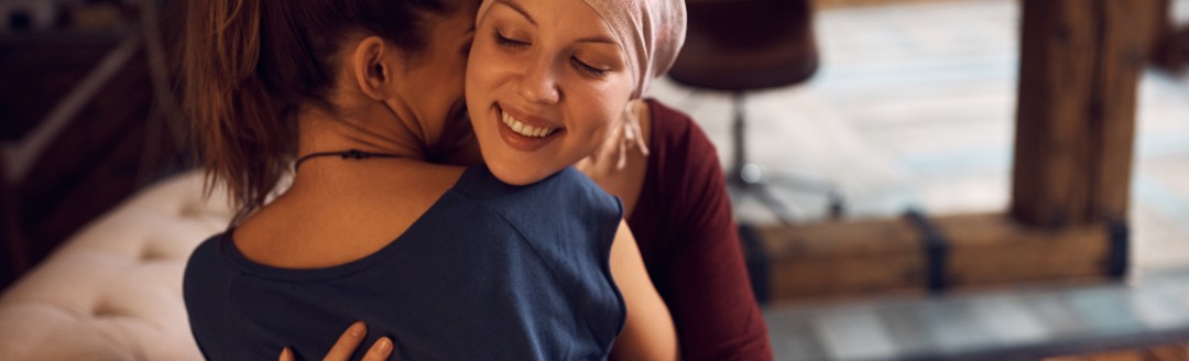 Zwei junge Frauen sitzen auf dem Sofa und umarmen sich. Eine der Frauen trägt hat ein Tuch auf dem Kopf.