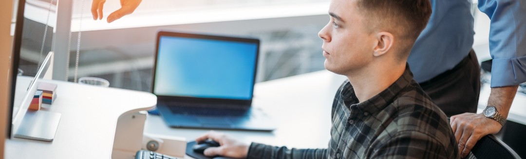 Ein junger Mann sitzt am PC und ein älterer Mann zeigt ihm etwas. 