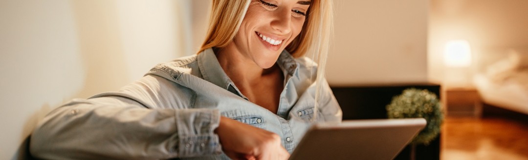 Eine junge Frau lächelt, während sie etwas auf ihr Tablet durchliest. Sie sitzt in ihrem Wohnzimmer auf einer Couch.