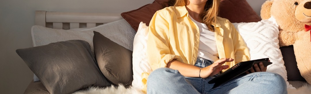 Eine junge Frau sitzt mit einem Riesen-Teddy auf ihrem Bett und hält ein Tablet in der Hand.