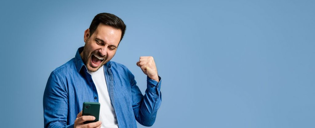 Mann mit weißem T-Shirt und blauem, offenen Hemd sieht auf sein Handy und freut sich