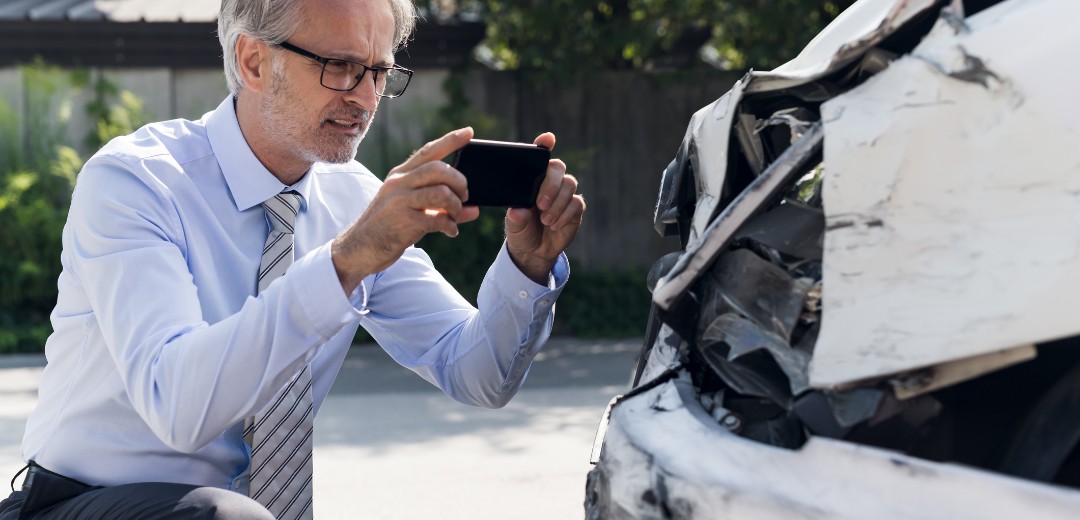 Ein älterer Mann fotografiert mit seinem Mobiltelefon sein Auto, dass einen Schaden erlitten hat.