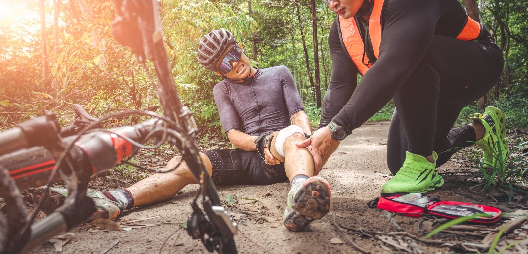 Ein Fahrradfahrer sitzt verletzt auf dem Waldboden. Sein Fahrrad liegt neben ihm und seine Verletzungen werden von einem Helfer angeschaut. 