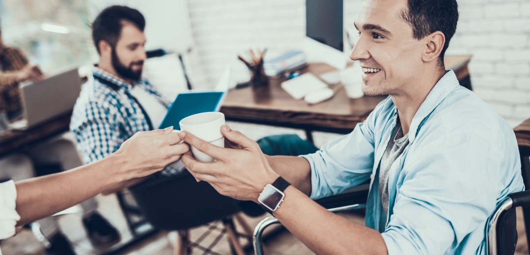 Ein junger Mann sitzt im Rollstuhl und ihm wird eine Tasse Kaffee gereicht. Mit ihm sitzt ein Kollege im Büro. 