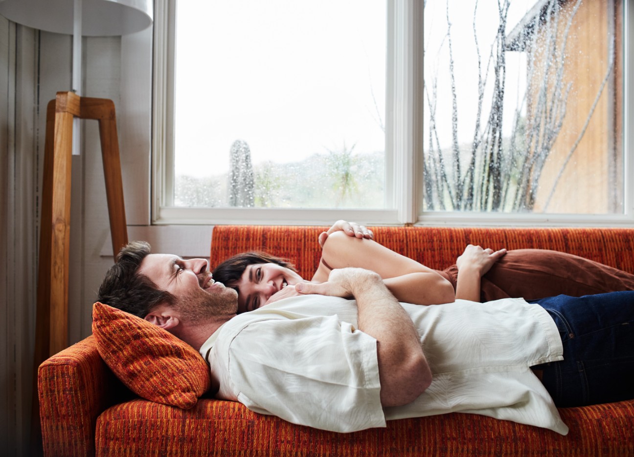Mann und Frau liegen aneinander gekuschelt auf einem Sofa während es draußen stark regnet