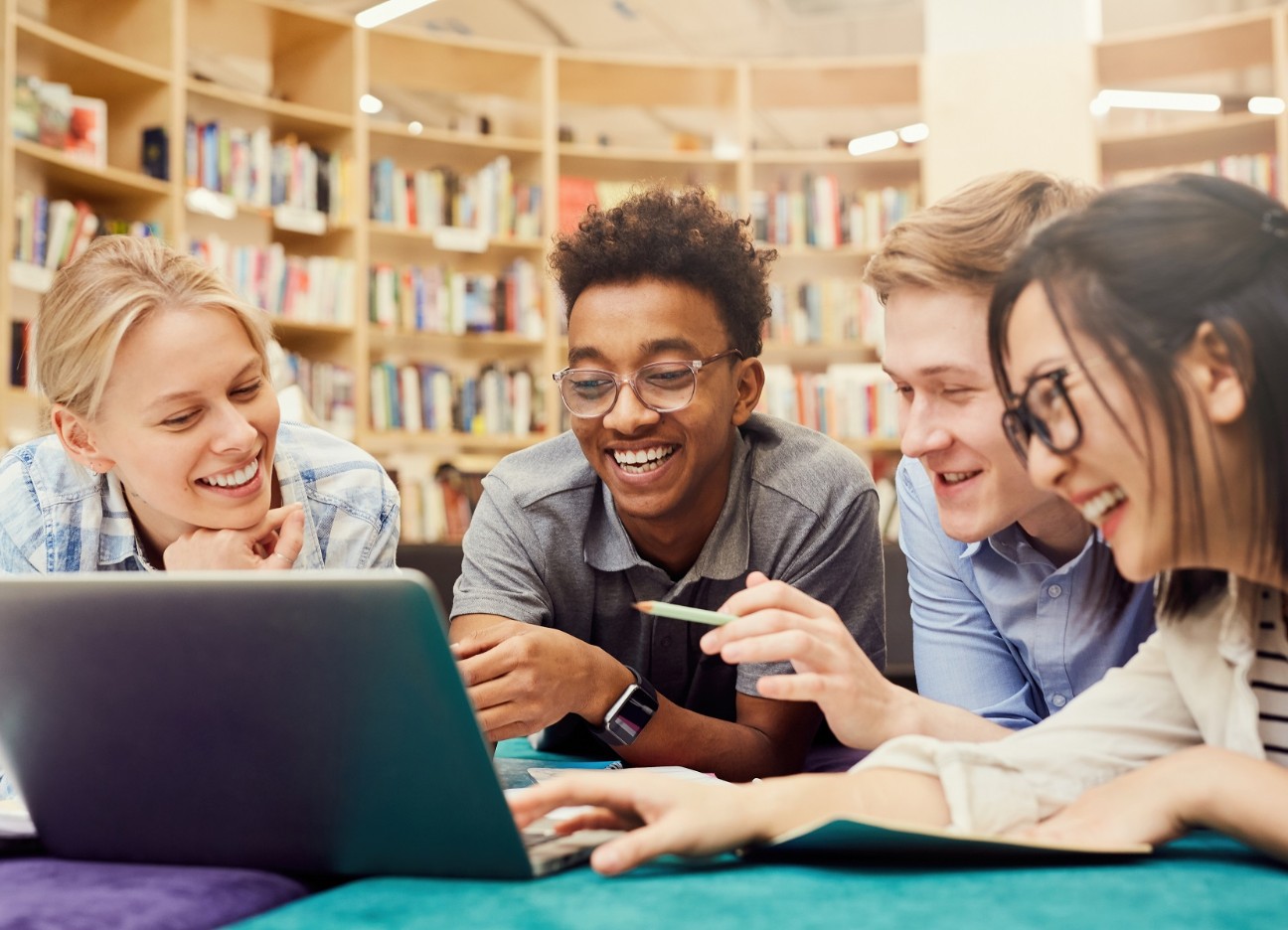 Gute gelaunte multiethnische Studenten in lässiger Kleidung schauen in der Campus Bibliothek ein Video auf dem Laptop an. 