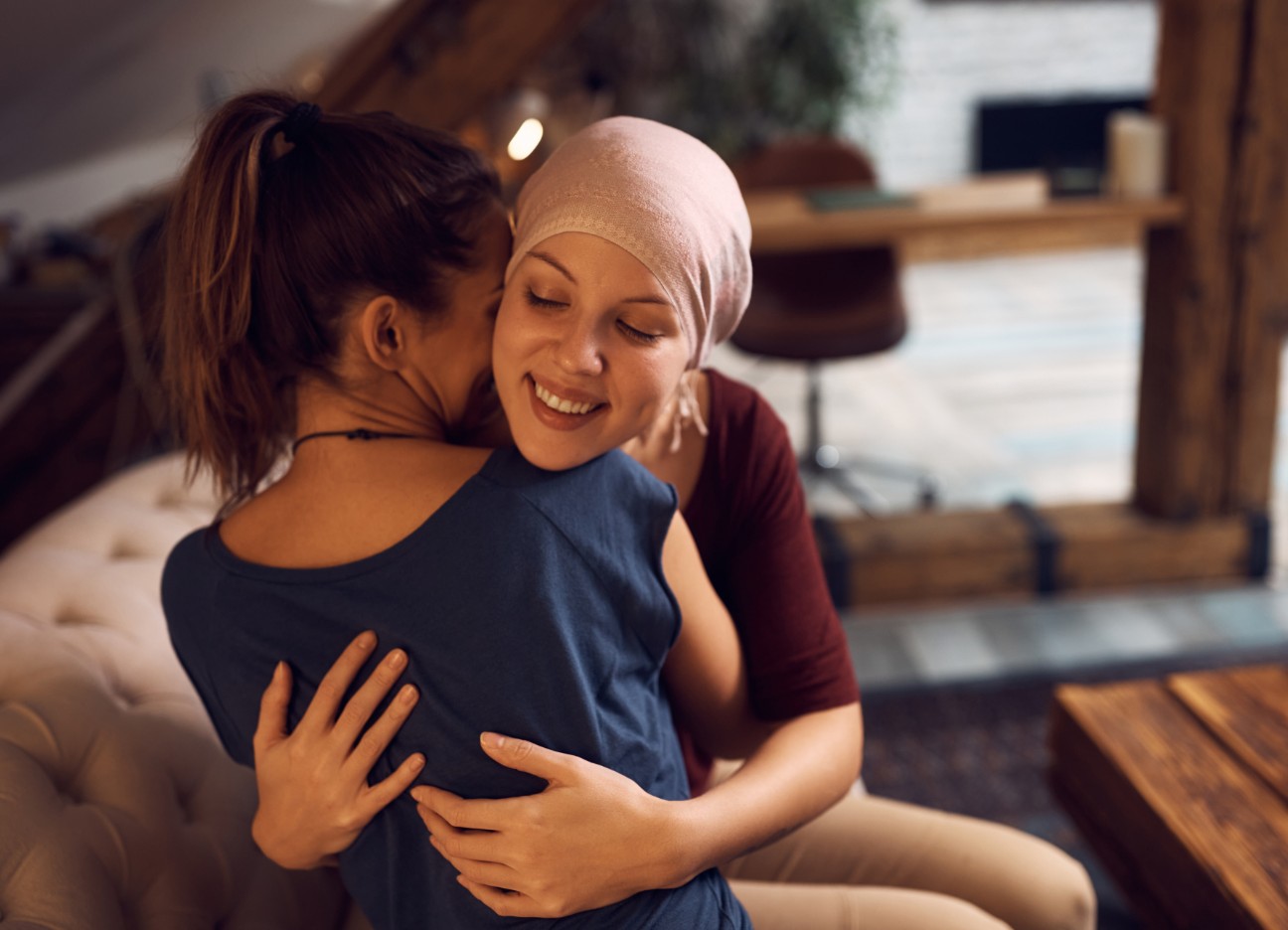 Zwei junge Frauen sitzen auf dem Sofa und umarmen sich. Eine der Frauen trägt hat ein Tuch auf dem Kopf.