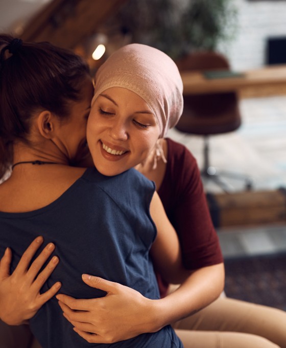 Zwei junge Frauen sitzen auf dem Sofa und umarmen sich. Eine der Frauen trägt hat ein Tuch auf dem Kopf.