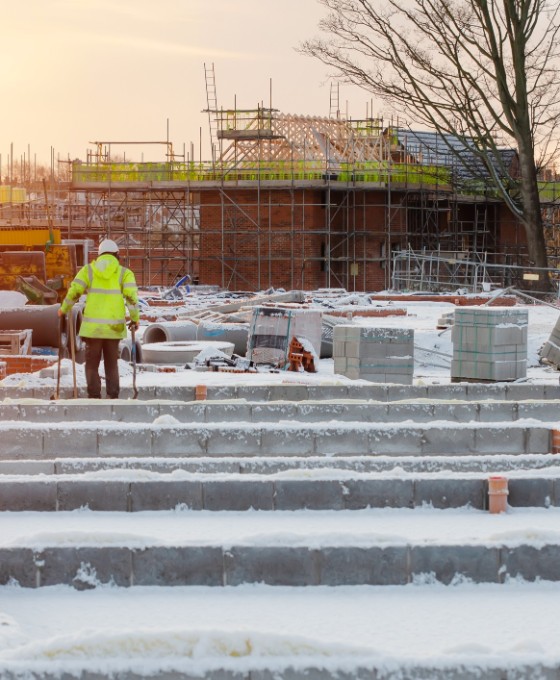 Dach einer verschneiten Baustelle. In Ferne kann ein Bauarbeiter in gelber Warn-Schutzkleidung erkannt werden. 