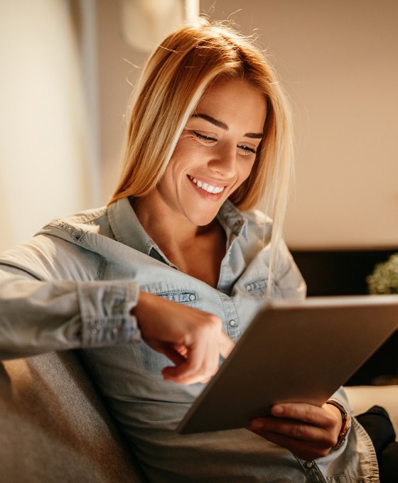 Eine junge Frau lächelt, während sie etwas auf ihr Tablet durchliest. Sie sitzt in ihrem Wohnzimmer auf einer Couch.