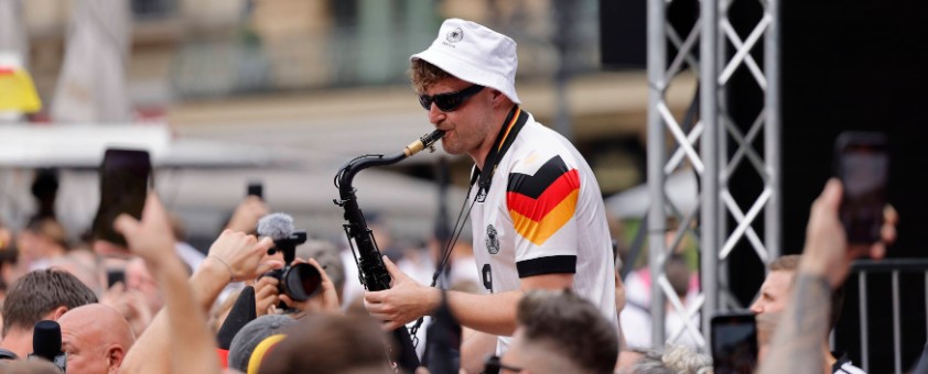 Andre Schnura, mit Deutschland-Trikot, spielt Saxophon in einer Menschenmenge von Fußballfans.