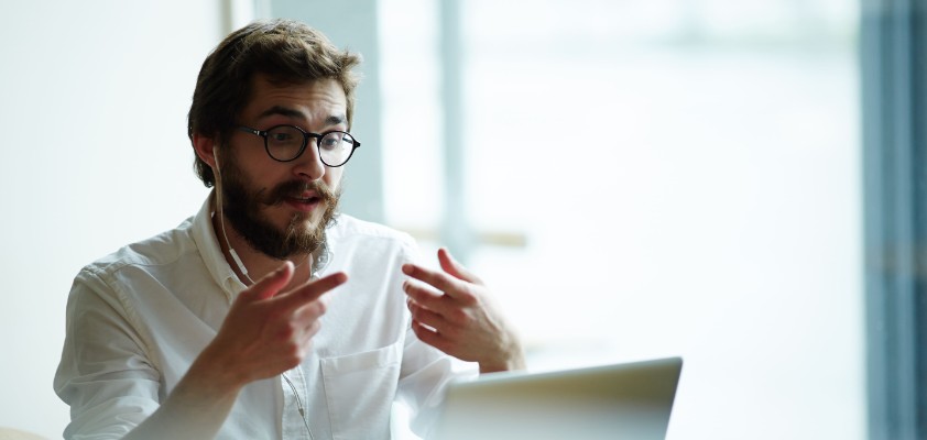 Ein Mann sitzt am Laptop in einem Online-Meeting und benutzt seine Hände für Gestiken. 