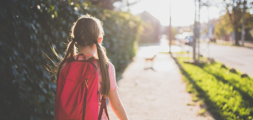 Ein junges Mädchen, fotografiert von hinten, läuft mit ihrem Rucksack eine Straße entlang.