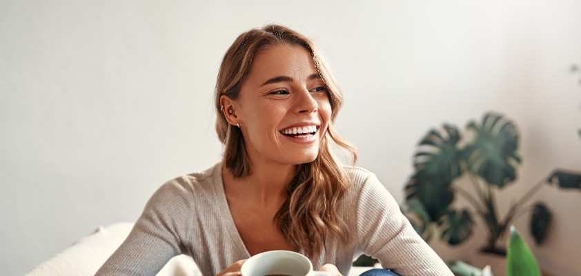 Eine Frau sitz lächelnd mit einer Tasse Kaffee auf dem Sofa