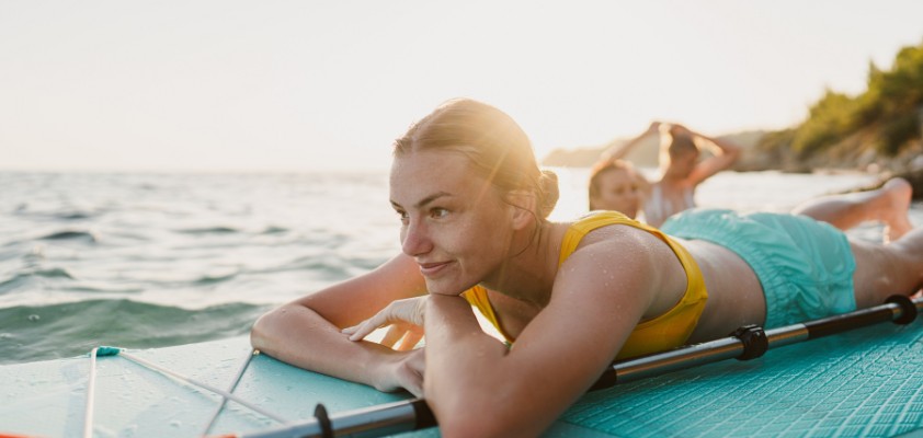 Eine junge Frau liegt auf dem Standup-Paddle-Board auf dem Wasser und schaut in die Ferne.