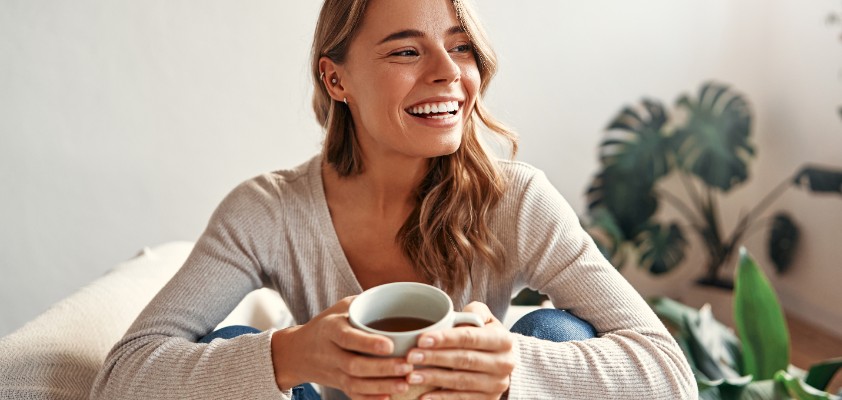 Eine Frau sitz lächelnd mit einer Tasse Kaffee auf dem Sofa