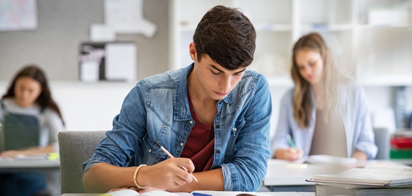 Ein Schüler der Sekundarstufe sitzt im Klassenzimmer an einem Tisch und schreibt etwas in seinen Block