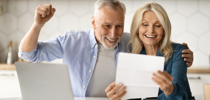 Ein älteres Paar freut sich über einen in der Hand haltenden Vertrag, während sie am Laptop in der Küche sitzen