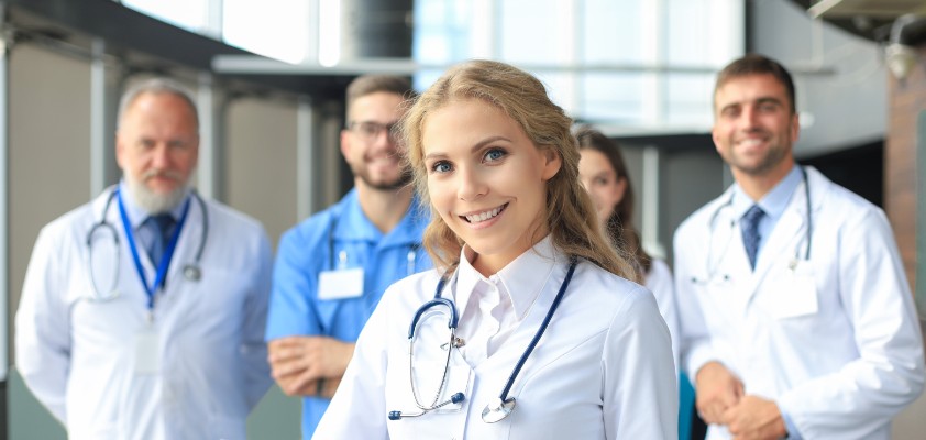 Eine Gruppe von Ärzten lächelt in die Kamera. Eine junge Ärztin steht im Vordergrund und hält ein Clipboard in der Hand.