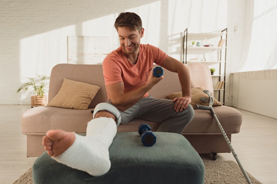 Ein Mann sitzt mit einem Gipsbein auf dem Sofa und verbildlicht, dass er mit Hanteln seine Arme trainiert. Er lächelt und schaut zu seinem Gipsbein hinunter.