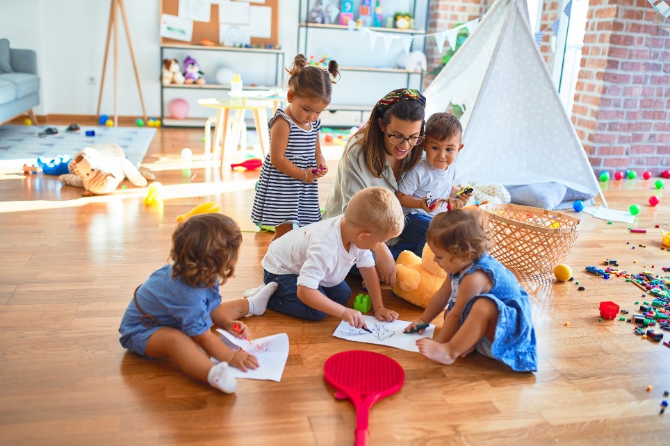 Schöne Lehrerin und Kleinkindgruppe sitzt mit Papier und Bleistift auf dem Boden und umspielt sich im Kindergarten viele Spielzeuge.