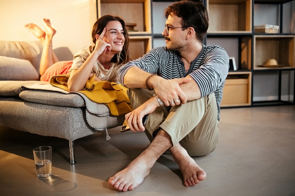 Junges Paar sitzt zufrieden im Wohnzimmer und lächelt sich an.