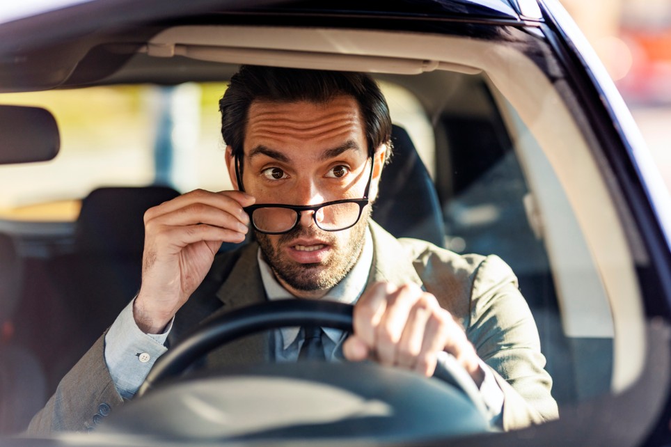 Ein Mann im Anzug sitzt im Auto am Lenker und schiebt überrascht seine Brille runter