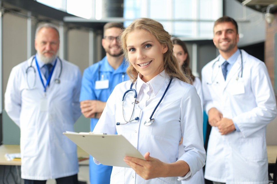 Eine Gruppe von Ärzten lächelt in die Kamera. Eine junge Ärztin steht im Vordergrund und hält ein Clipboard in der Hand.