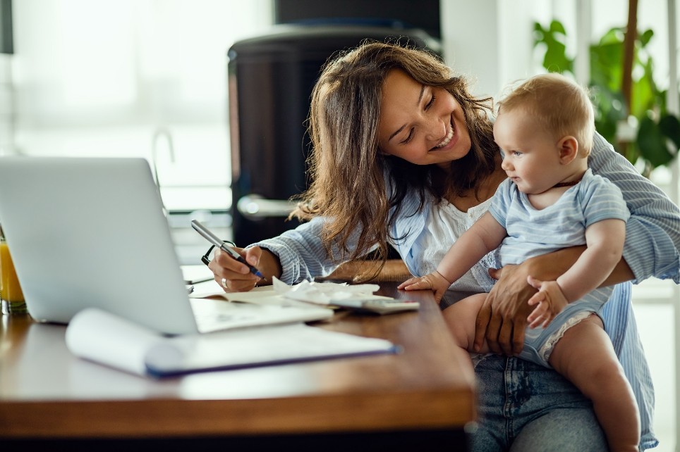 Glückliche Mutter spricht mit ihrem Baby während der Arbeit am Laptop zuhause. 