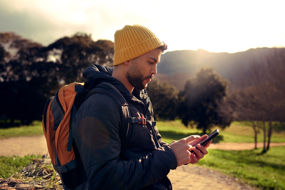 Junger Mann in Outdoor-Kleidung ist beim Wandern in den Bergen und schaut auf sein Handy